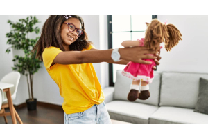 A girl holding a plush doll in the middle of her living room. She is wearing a yellow shirt and faded jeans.
