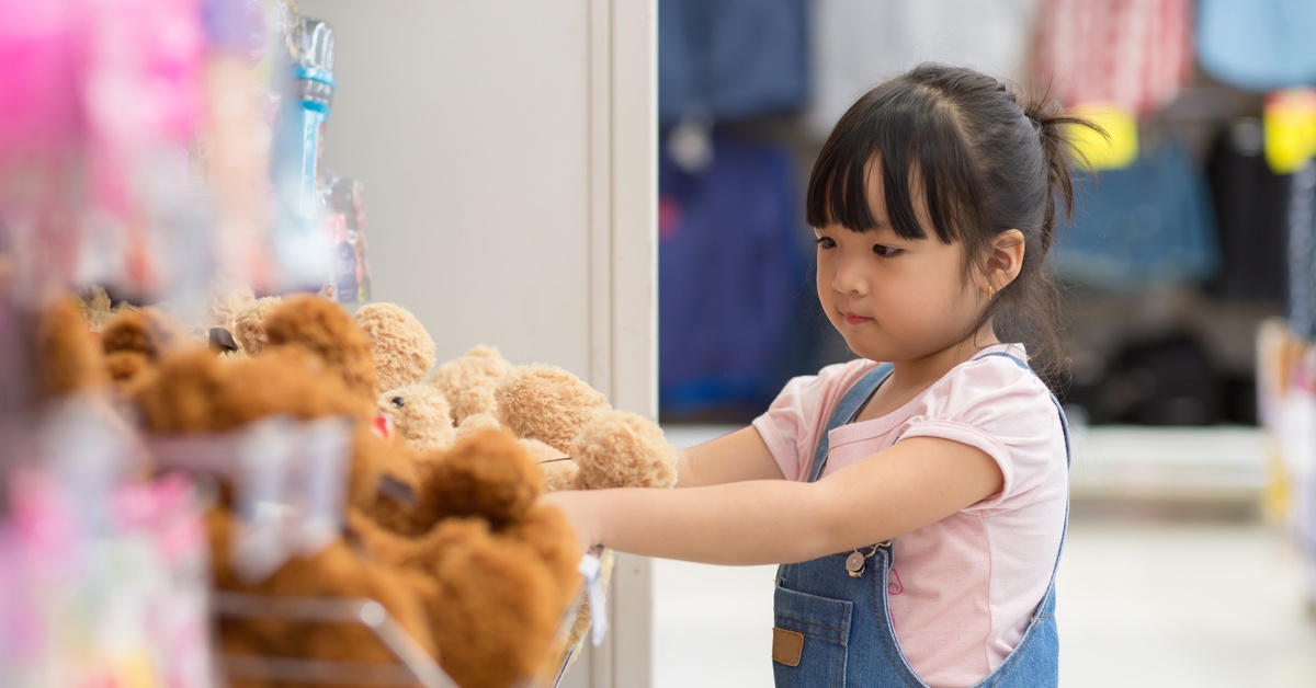 A little girl placing her hands on plushies inside of a retail store. She is wearing a pink shirt and overalls.