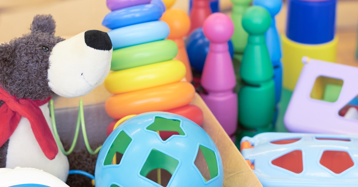 A pile of toys sitting in a cardboard box, including a plushie and a ball. There are more plastic toys outside of the box.