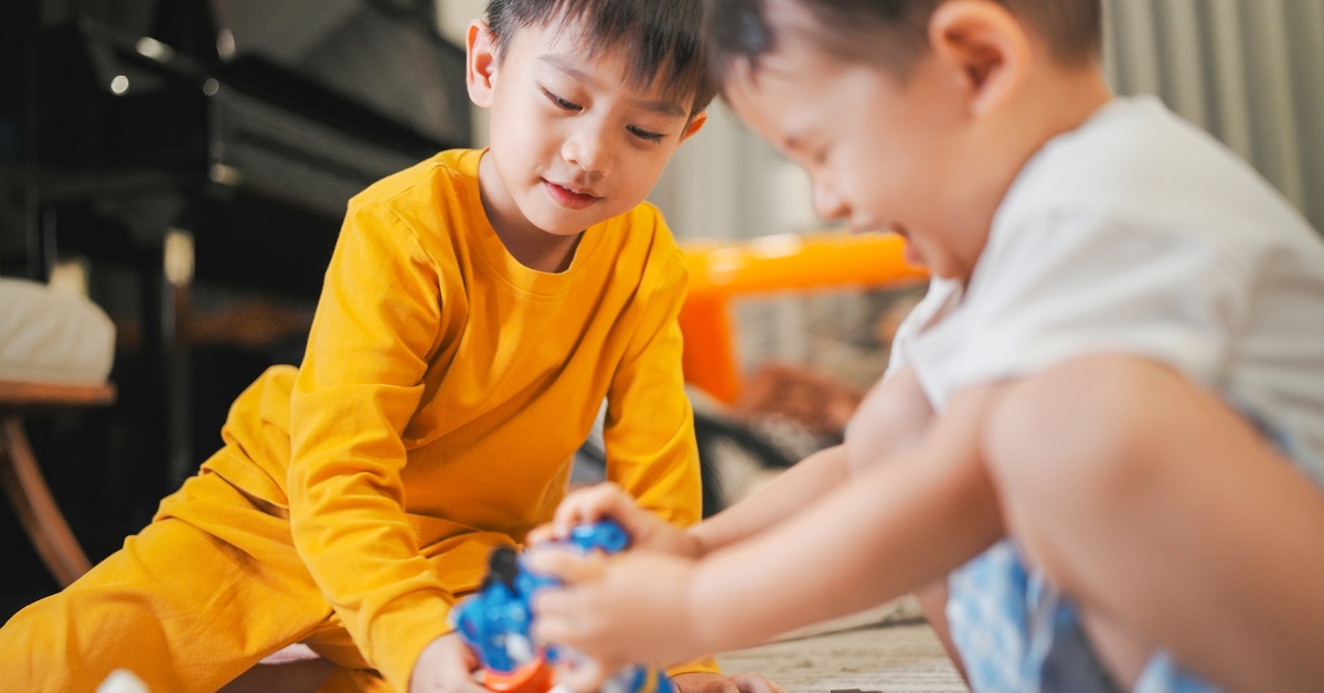 Two young boys playing with action figures on the floor of their home. One of them holds a blue, bulky toy.