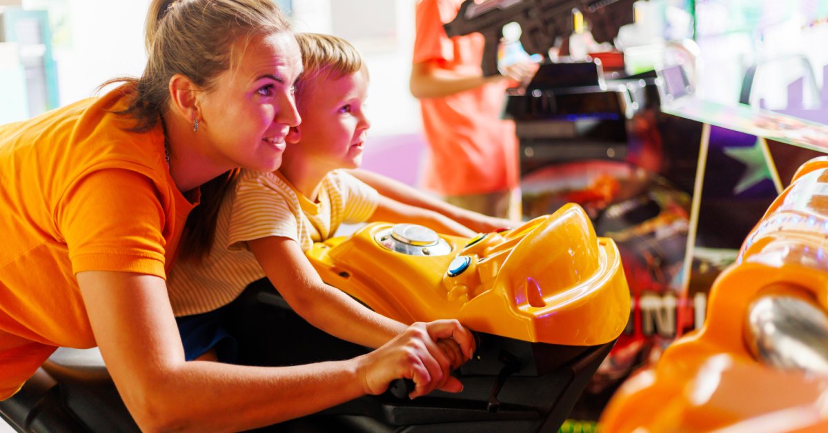 A mother and her son playing in an arcade. The son sits on a motorcycle machine while the mother controls the bike.