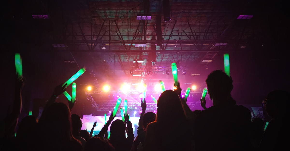A large crowd of people at a dark indoor event. They are holding green light sticks in the air facing the main stage.