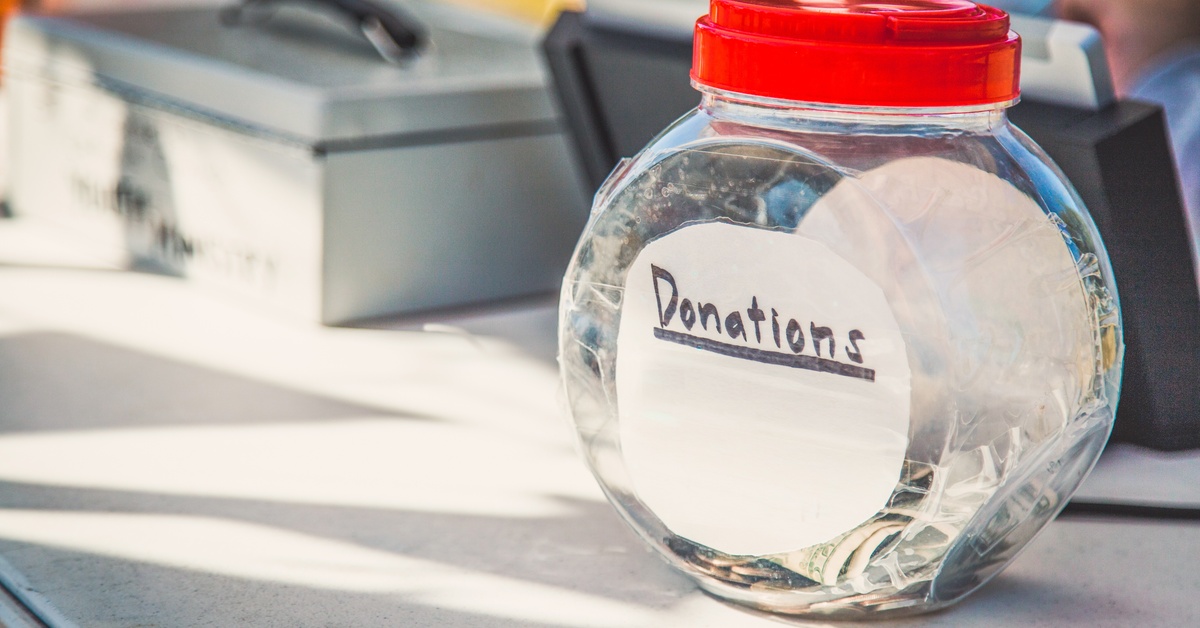 A container labeled “donations” filled with dollar bills and coins. It is sitting on a white table in front of metal boxes.