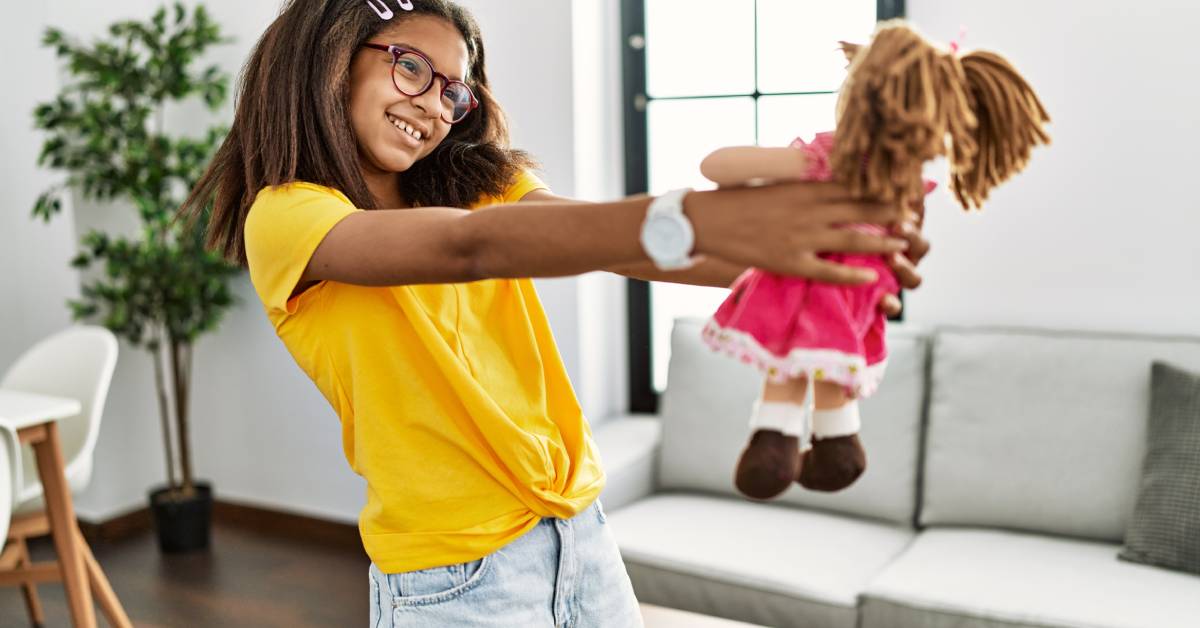 A girl holding a plush doll in the middle of her living room. She is wearing a yellow shirt and faded jeans.