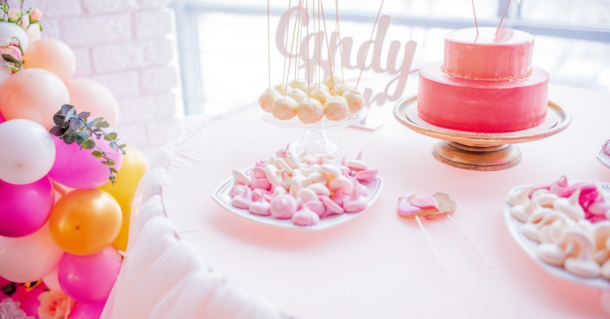 Pink and white birthday decorations set on a table. There are balloons, candy, and baked goods on display.