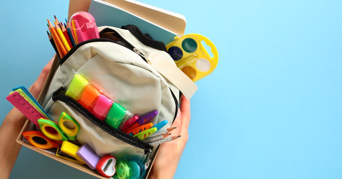 Two hands holding a box of school supplies. A backpack stuffed with writing utensils and other supplies sits in the box.