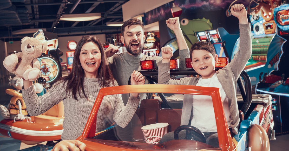 A mom, dad, and son at an indoor arcade. They raise their arms in the air as their son sits on an arcade car machine.