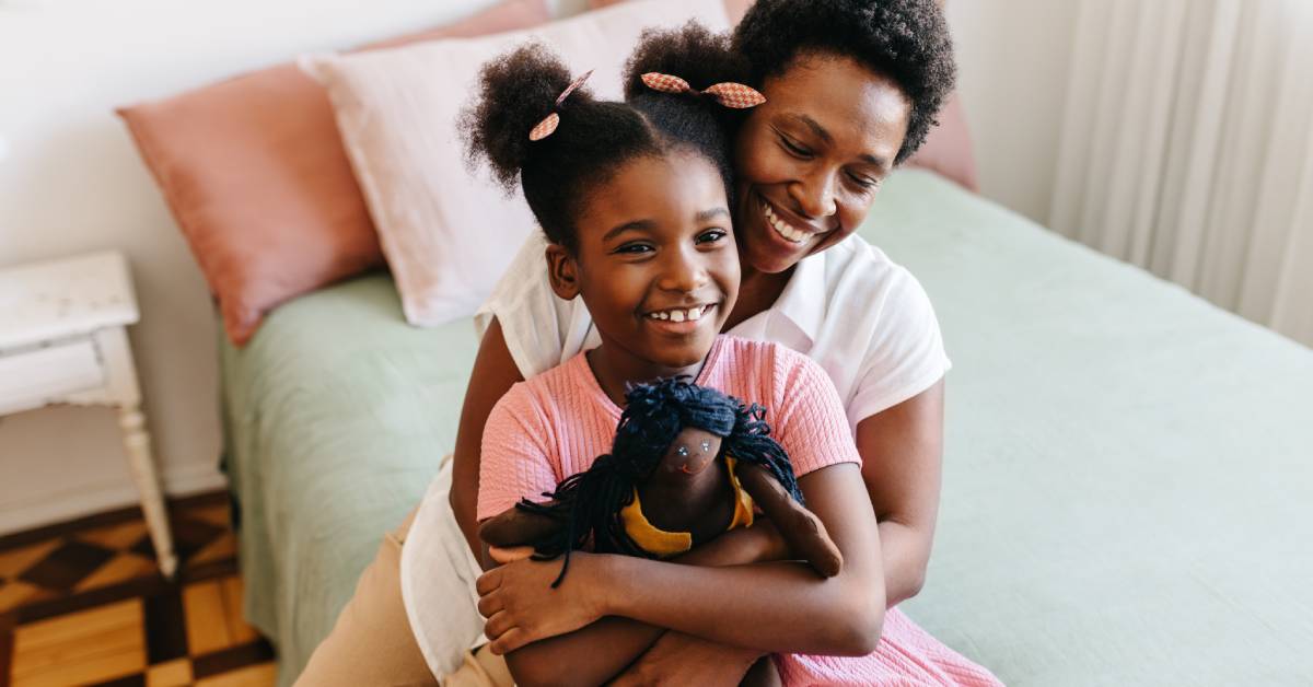 A mother and her daughter hug while they sit on a twin-sized bed. The daughter has a cloth doll in her arms.